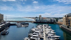A marina with boats docked, a clear sky, and buildings in the background. - Watercolours on Marina (Glenelg)