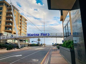 A pedestrian bridge with "Marina Pier" signage, a road with directional arrows, and a building with "Light's Landing Chapel Drive" signage. - Watercolours on Marina (Glenelg)