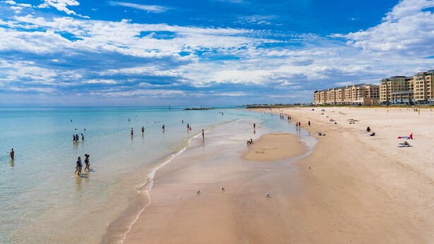 A beach with clear blue water, a sandy shore, and buildings in the background. - Watercolours on Marina (Glenelg)