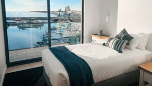 A bedroom with a large window, a bed with white bedding, a bedside table with a plant, and a view of a marina. - Watercolours on Marina (Glenelg)