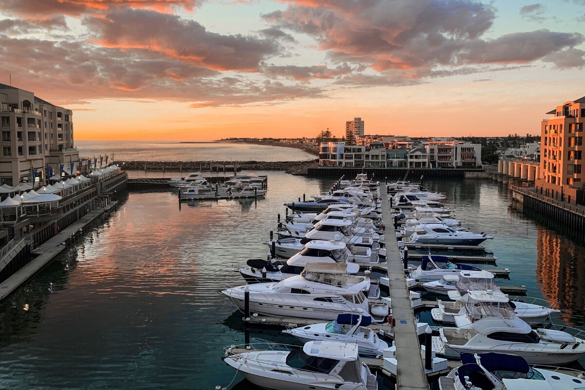 Un puerto deportivo con varias embarcaciones amarradas, un muelle que se adentra en el agua y edificios a lo largo del paseo marítimo.
