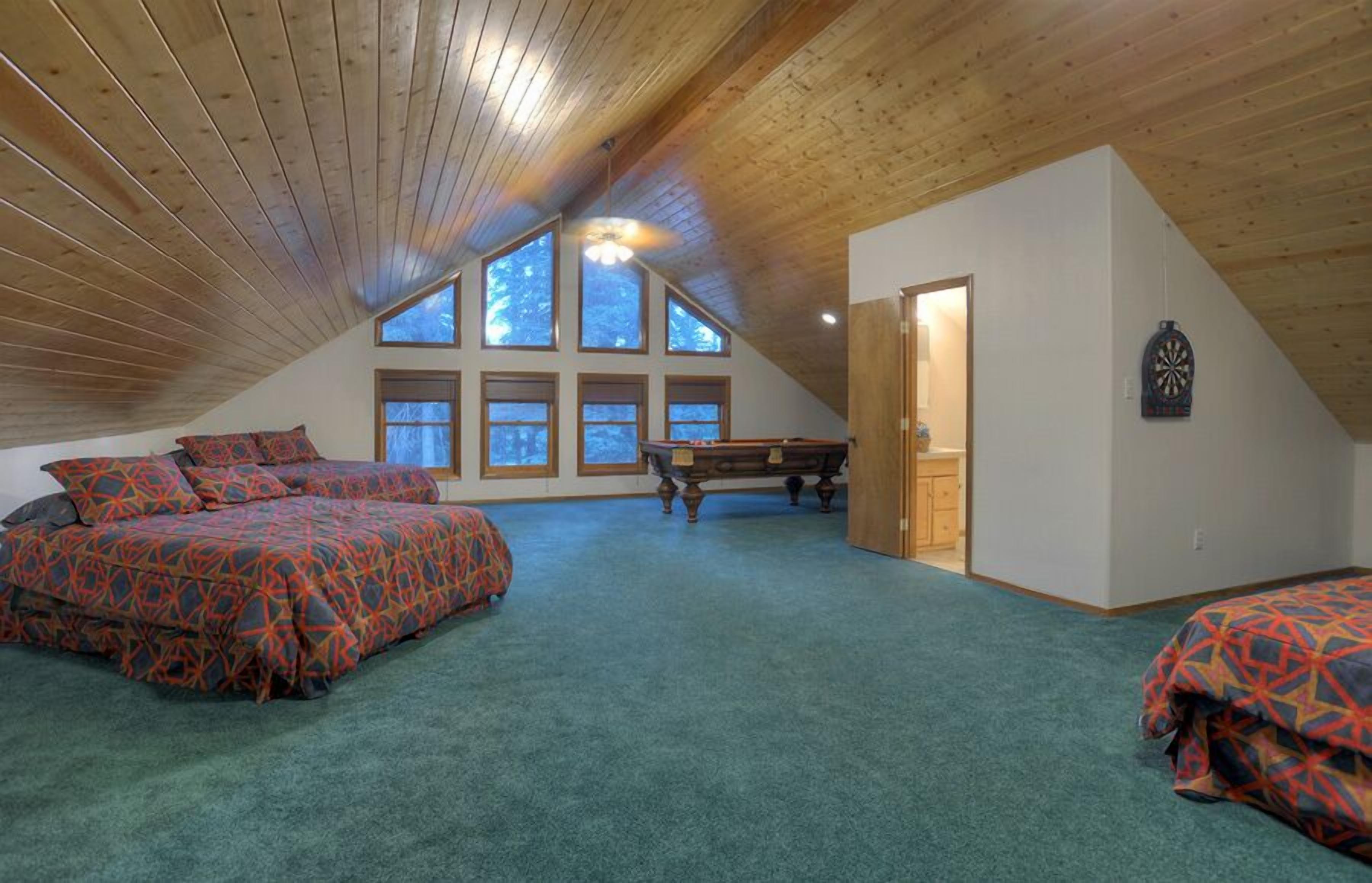 A spacious attic bedroom with a sloped ceiling, large windows, a pool table, and a dartboard.