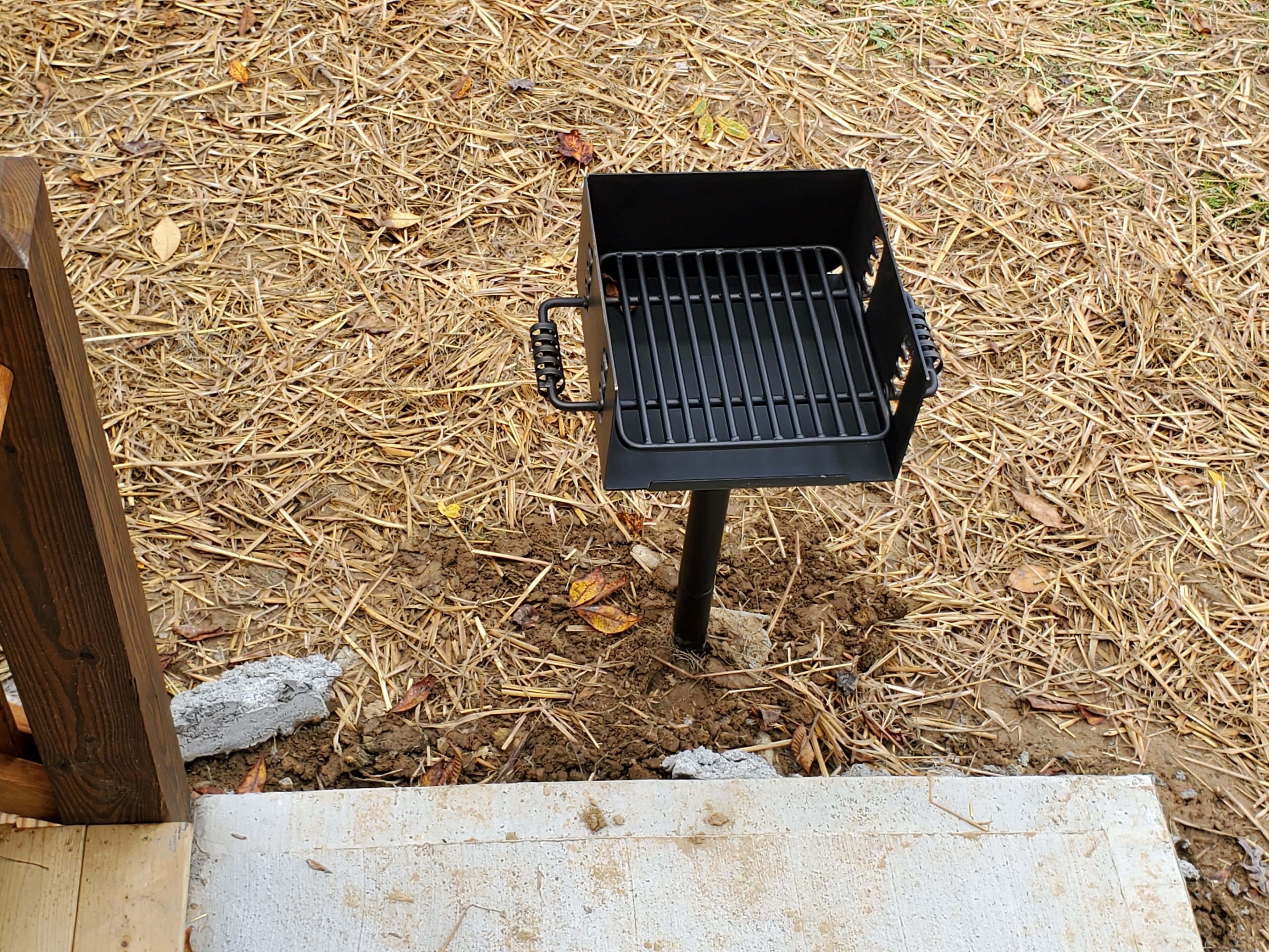 Un barbecue noir, dot é d’une grille en métal, est posé sur une terrasse en bois bordée de blanc, et est entouré de paillis et d’aiguilles de pin.