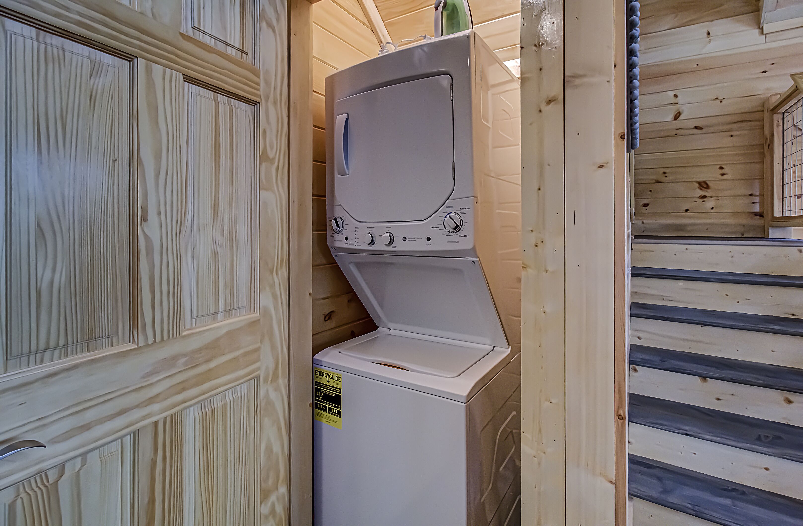 A compact, white, front-loading washing machine installed in a wooden cabin with a small window and a door with a decorative panel.