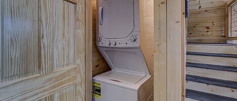 A compact, white, front-loading washing machine installed in a wooden cabin with a small window and a door with a decorative panel.