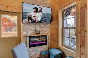 A cozy cabin interior with a mounted TV, a stone fireplace, and a large window overlooking a deck.