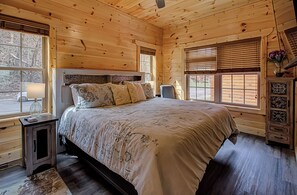 A wooden cabin bedroom with a large bed, a nightstand, a dresser, and a window with blinds.