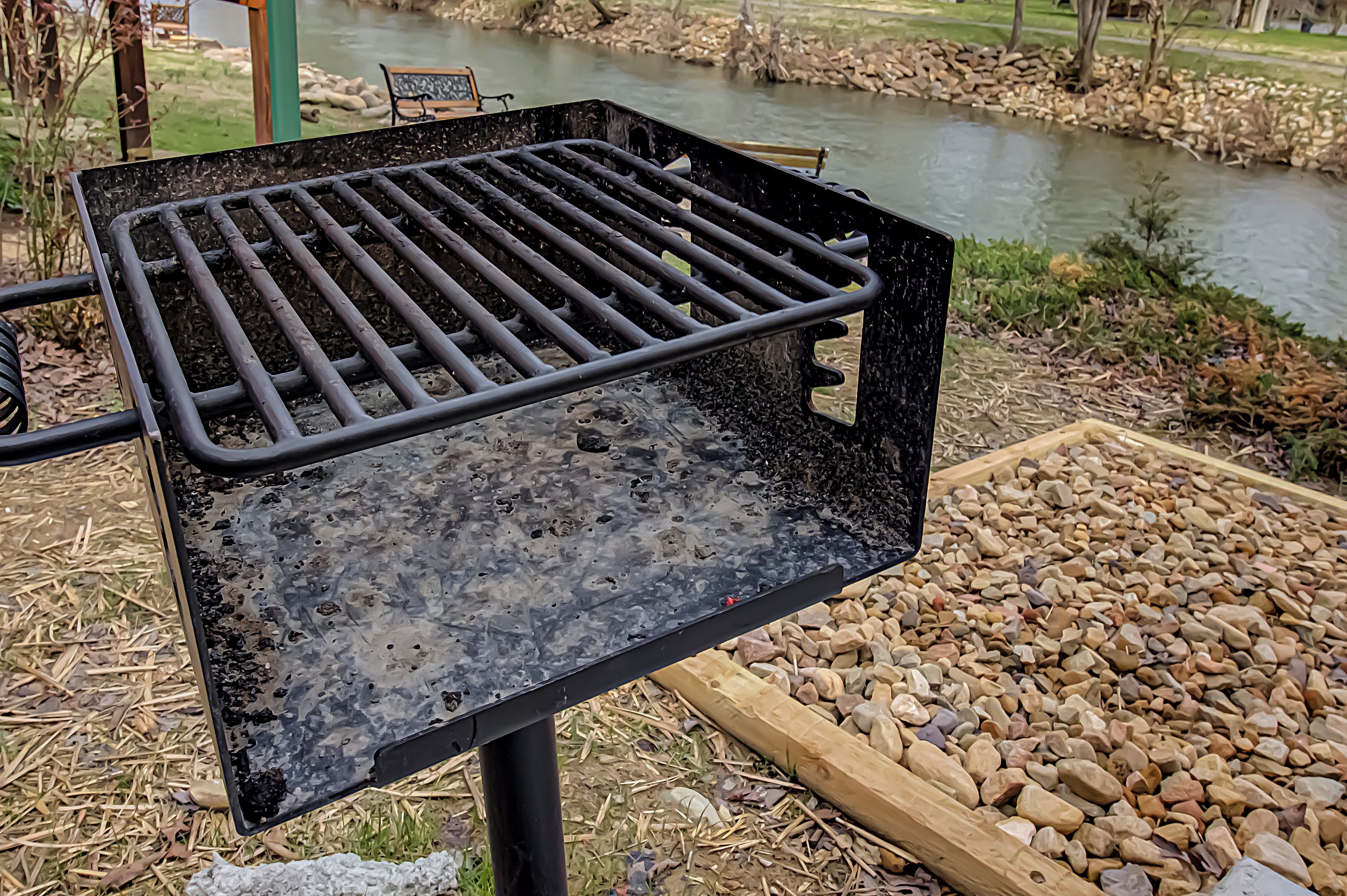 A grill with a metal grate, set against a backdrop of a river and pebbles.