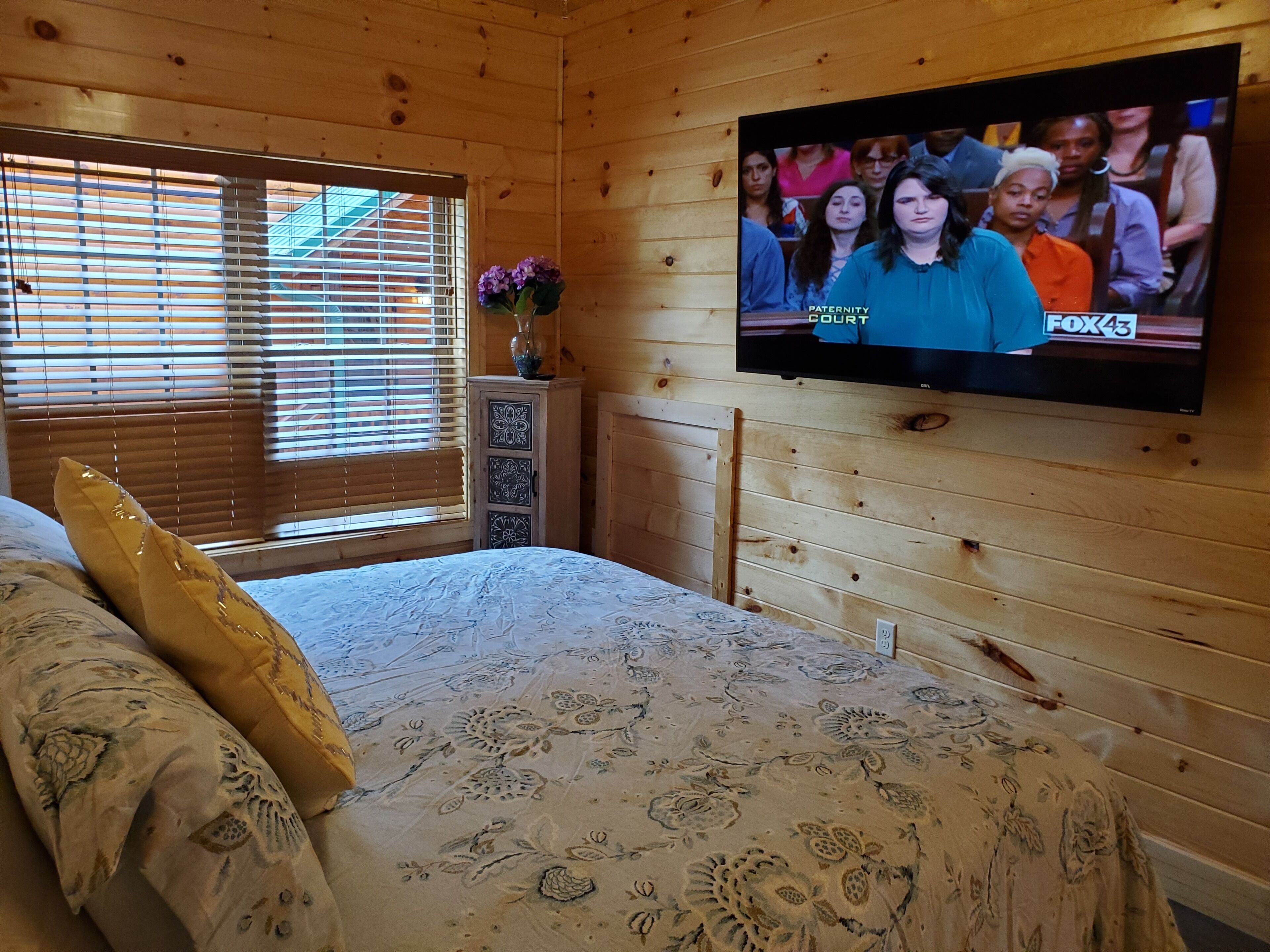 A wooden cabin room with a bed, a TV mounted on the wall, and a window with blinds.