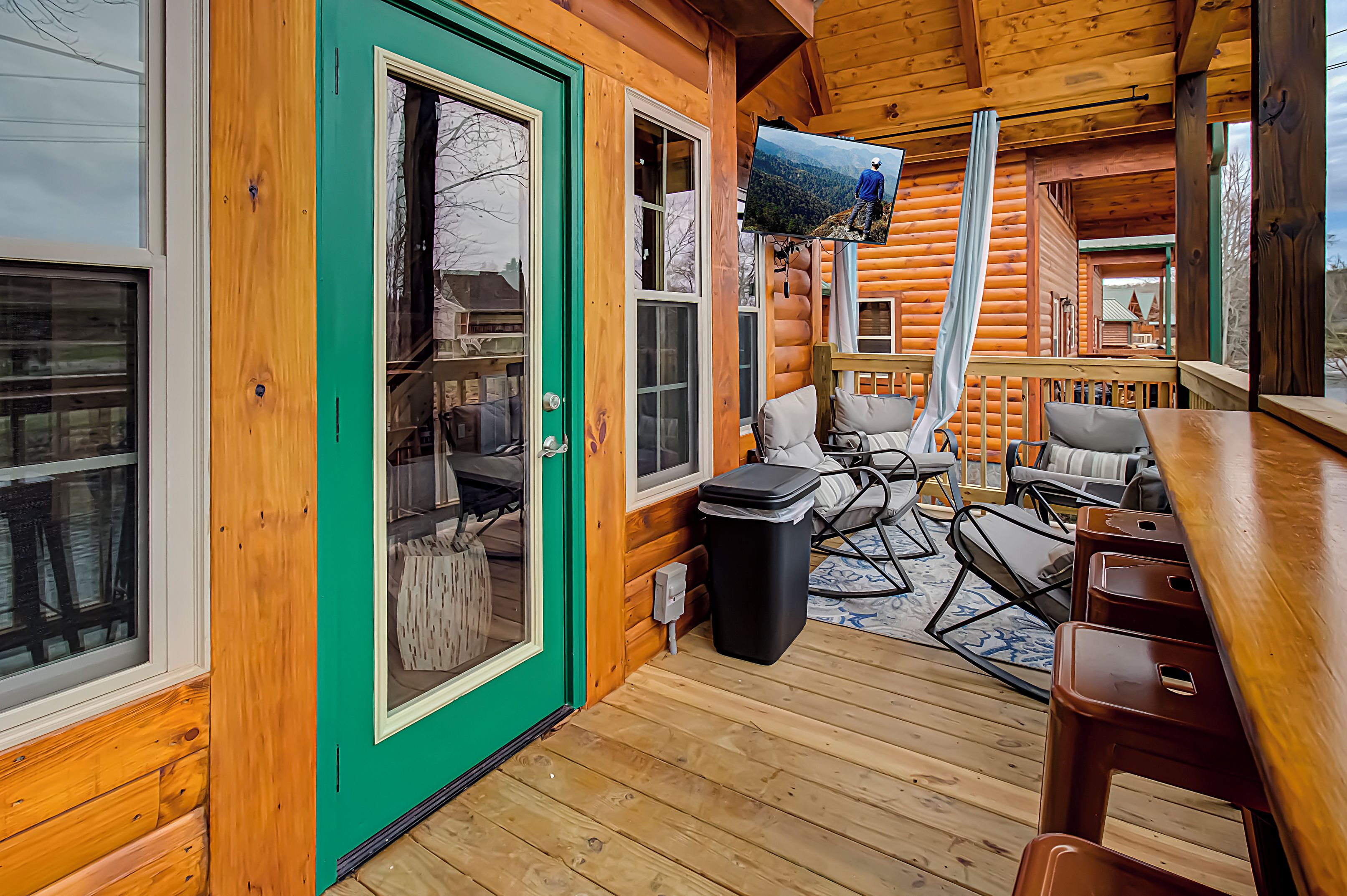 Une cabane en rondins avec une porte verte, une terrasse en bois, un téléviseur fixé au mur et un coin salon avec des chaises et une table.