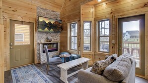 A cozy cabin interior with a stone wall, wooden floors, a fireplace, a sofa, a bench, and large windows offering views of the outside. - River View | Pigeon Forge  | Arcade | Hot Tub | WiFi | Fireplace (Pigeon Forge)