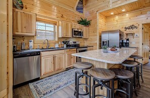 A wooden cabin-style kitchen with a central island, bar stools, and a 'gather' sign on the wall.
