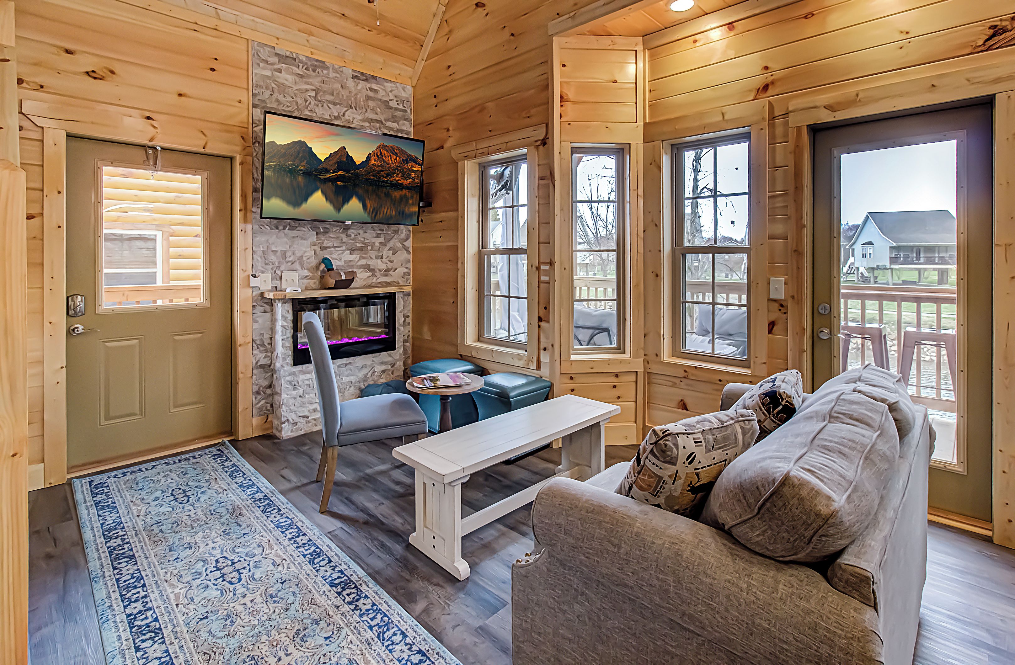 Interior acogedor de una cabaña con pared de piedra, piso de madera, chimenea, sofá, banco y ventanas grandes que ofrecen vistas al exterior.