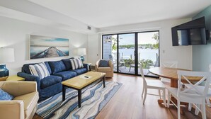 A modern living room with a sofa, coffee table, and chairs, a dining table with chairs, a TV mounted on the wall, and a large window overlooking a body of water.