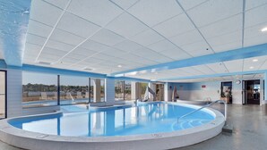 Une grande piscine intérieure à l’eau d’un bleu limpide, entourée d’une terrasse carrelée et d’un espace abrité avec des chaises longues.