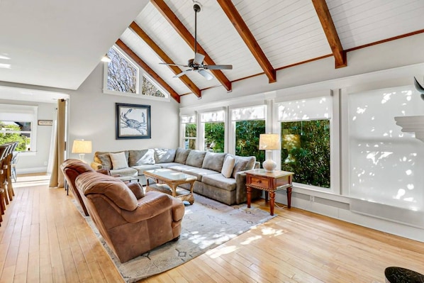 A spacious living room with a high ceiling, wooden beams, a large sofa set, a coffee table, and a view of greenery outside.