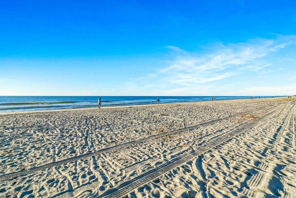 A sandy beach with tire tracks, a clear blue sky, and the ocean in the background.