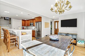 A modern kitchen with wooden cabinets, stainless steel appliances, a dining area with a wooden table, and a chandelier.