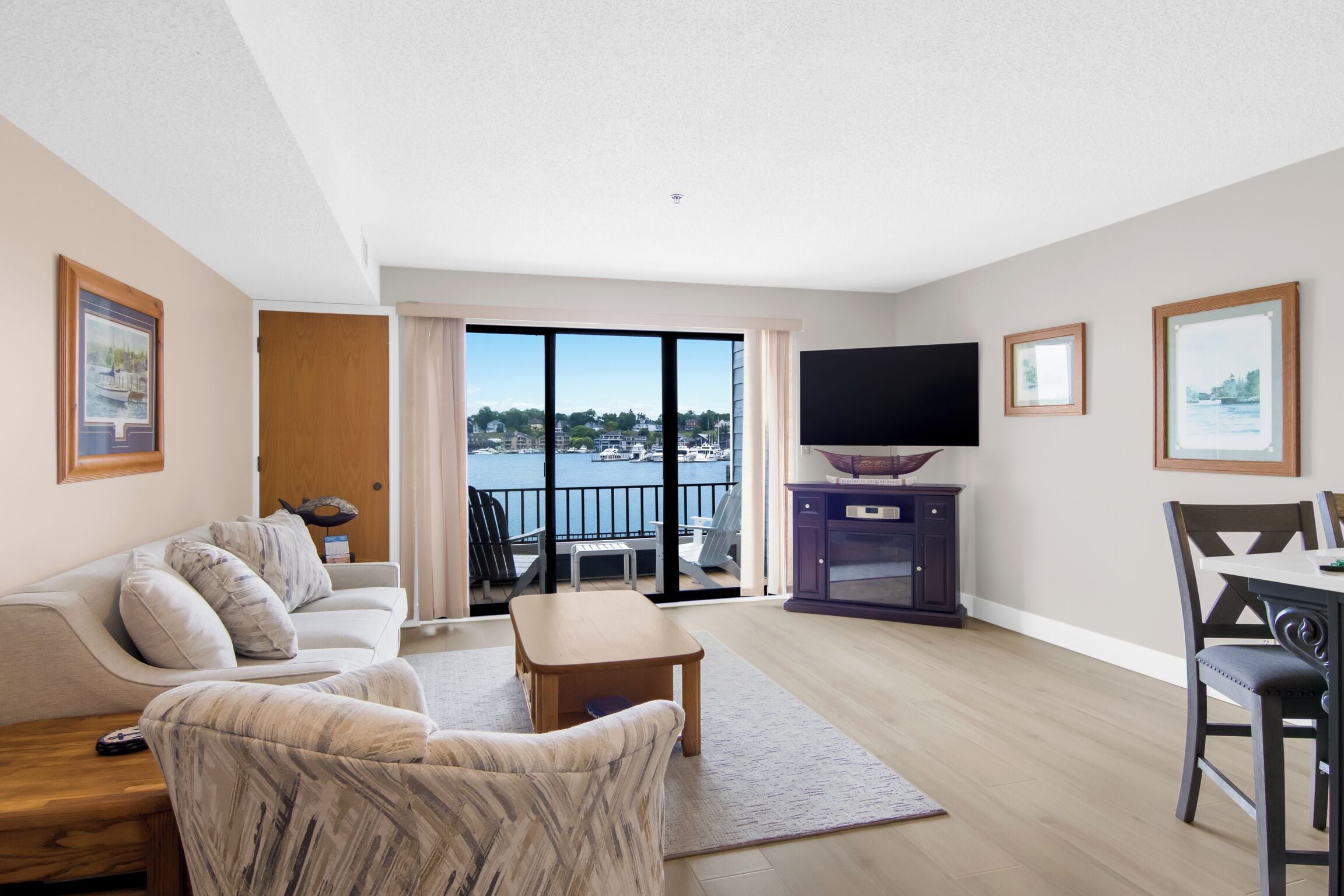 A modern living room with a sofa, a coffee table, and a TV. There is a balcony with a view of the water.