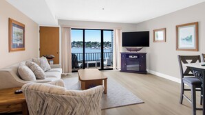 A modern living room with a sofa, a coffee table, and a TV. There is a balcony with a view of the water. - 112 EWI Lake Charlevoix View Condo | Pool, Hot Tub, Sauna | Fireplace (Charlevoix)