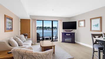A modern living room with a sofa, a coffee table, and a TV. There is a balcony with a view of the water.