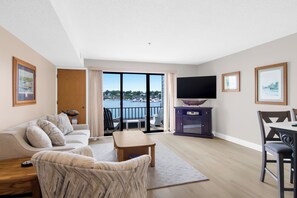 A modern living room with a sofa, a coffee table, and a TV. There is a balcony with a view of the water. - 112 EWI Lake Charlevoix View Condo | Pool, Hot Tub, Sauna | Fireplace (Charlevoix)