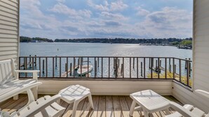 A wooden deck with white plastic chairs and a table, overlooking a calm body of water with boats and a distant shoreline. - EWI 222 | Downtown Charlevoix | Pool, Hot Tub, Sauna (Charlevoix)