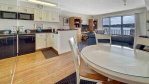 A modern kitchen with white cabinets, a dining area with a glass table, and a view of a body of water through large windows. - EWI 222 | Downtown Charlevoix | Pool, Hot Tub, Sauna (Charlevoix)