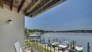 A balcony with white chairs, a view of a calm lake with boats, and a dock area. - 223 Edgewater Inn | Pool, Hot Tub, Sauna | Cozy 1 BR Condo (Charlevoix)