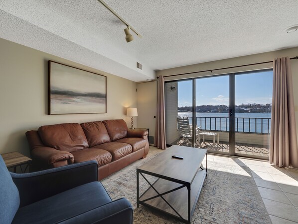 A modern living room with a brown leather sofa, a blue armchair, a wooden coffee table, and a large window offering a view of a body of water and buildings.