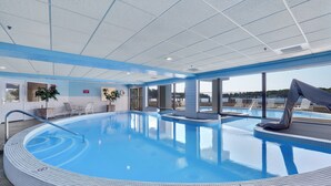 Une grande piscine intérieure à l’eau d’un bleu limpide, entourée d’une terrasse carrelée et d’un espace abrité avec des chaises longues.