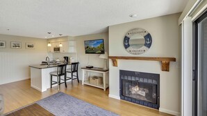 A modern kitchen with white cabinets, a dining area with two chairs, a fireplace, and a mounted TV. - 235 Edgewater | Scenic One-BDRM | Downtown Charlevoix | Pool, Hot Tub, Sauna (Charlevoix)