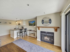 A modern kitchen with white cabinets, a dining area with two chairs, a fireplace, and a mounted TV. - 235 Edgewater | Scenic One-BDRM | Downtown Charlevoix | Pool, Hot Tub, Sauna (Charlevoix)