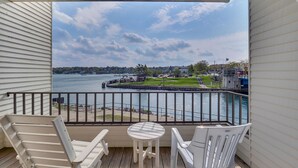 A balcony with a view of a lake, a dock, and a grassy area with a small building. - 239 Edgewater | Waterfront Views | Downtown Charlevoix | Pool, Hot Tub, Sauna (Charlevoix)