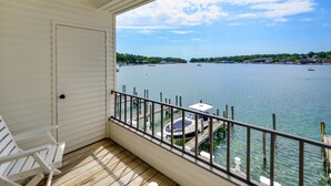 Un balcon offrant une vue sur un lac paisible, des bateaux amarrés et une côte lointaine parsemée de bâtiments.