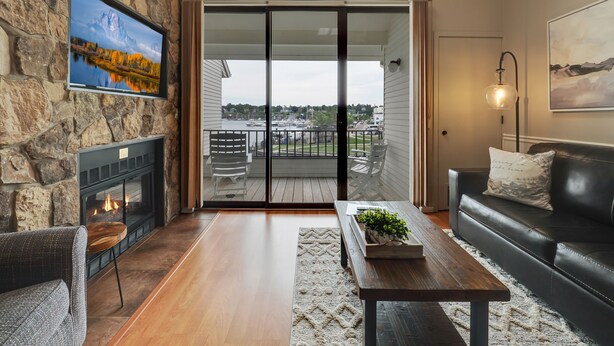 A living room with a stone wall, a fireplace, a TV, a balcony with a view of a marina, and a cozy seating area. - Edgewater 357 | Downtown Charlevoix | Pool, Hot Tub, Sauna (Charlevoix)