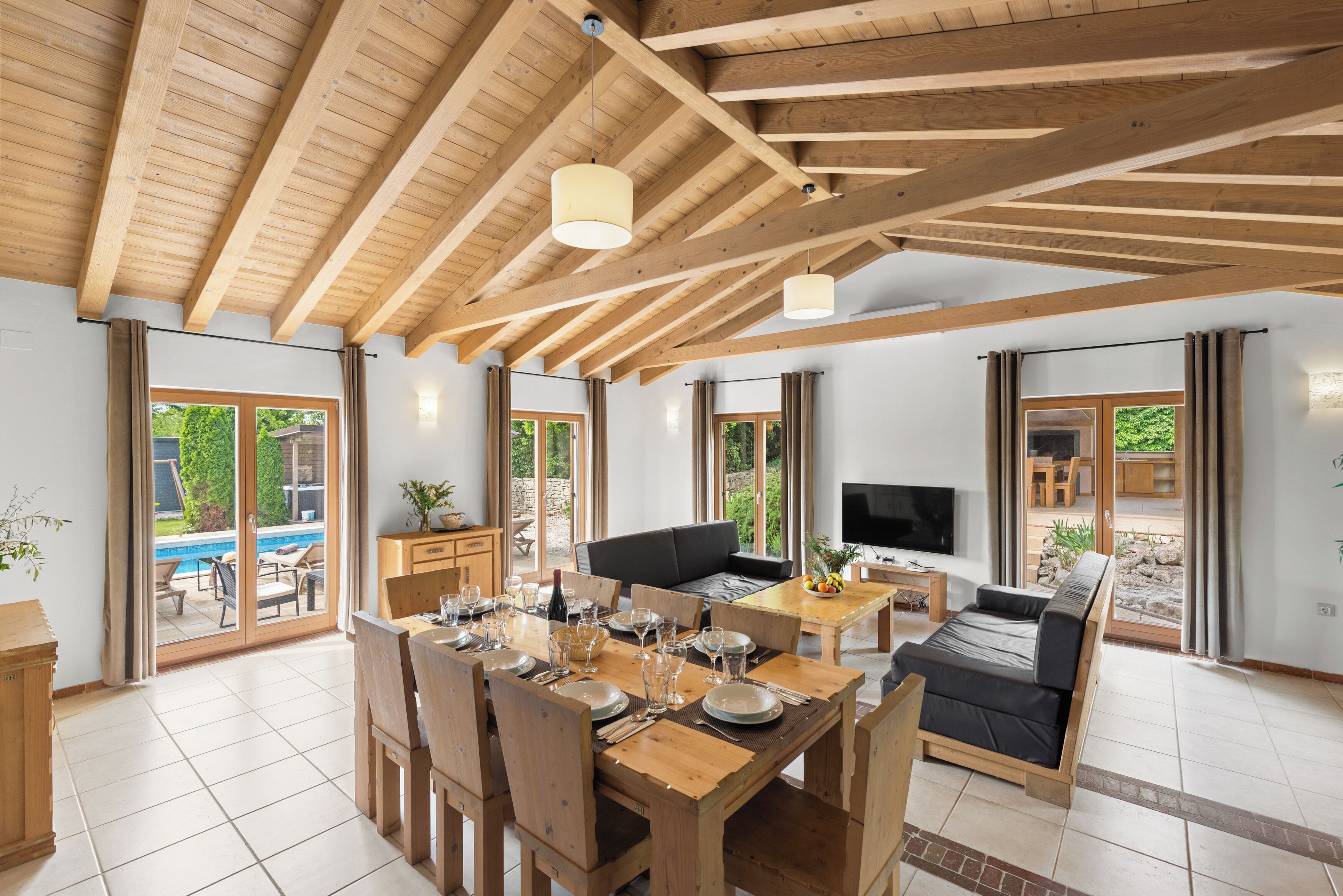 Amplia sala de estar con techo de madera y ventanas grandes, una mesa de comedor preparada para seis personas y un sofá.