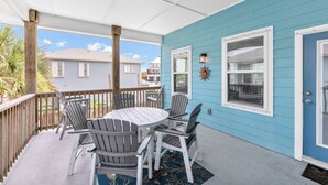 Un patio coperto con un tavolo rotondo e poltrone Adirondack, ringhiera in legno e parete esterna blu.