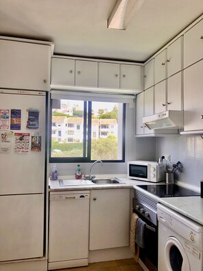 Cozinha compacta com armários brancos, micro-ondas embutido, máquina de lavar e janela com vista para edifícios.