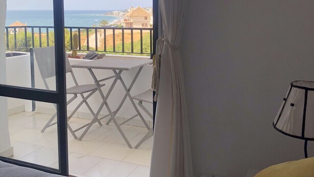 A room with a large sliding glass door leading to an outdoor balcony with a view of the sea and a building.