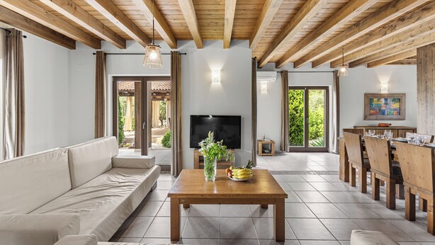 A spacious living area with a wooden ceiling, a large flat-screen TV, a sofa, a dining table with chairs, and a view of the outdoors.