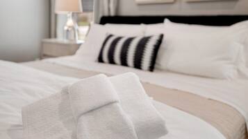 Una cama bien hecha con sábanas blancas y un cojín a rayas, una lámpara de noche y cuadros en la pared.