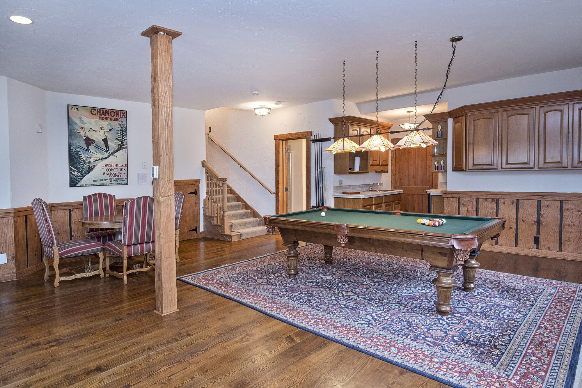 A pool table in a room with wooden flooring, a patterned rug, and a staircase leading to another area.