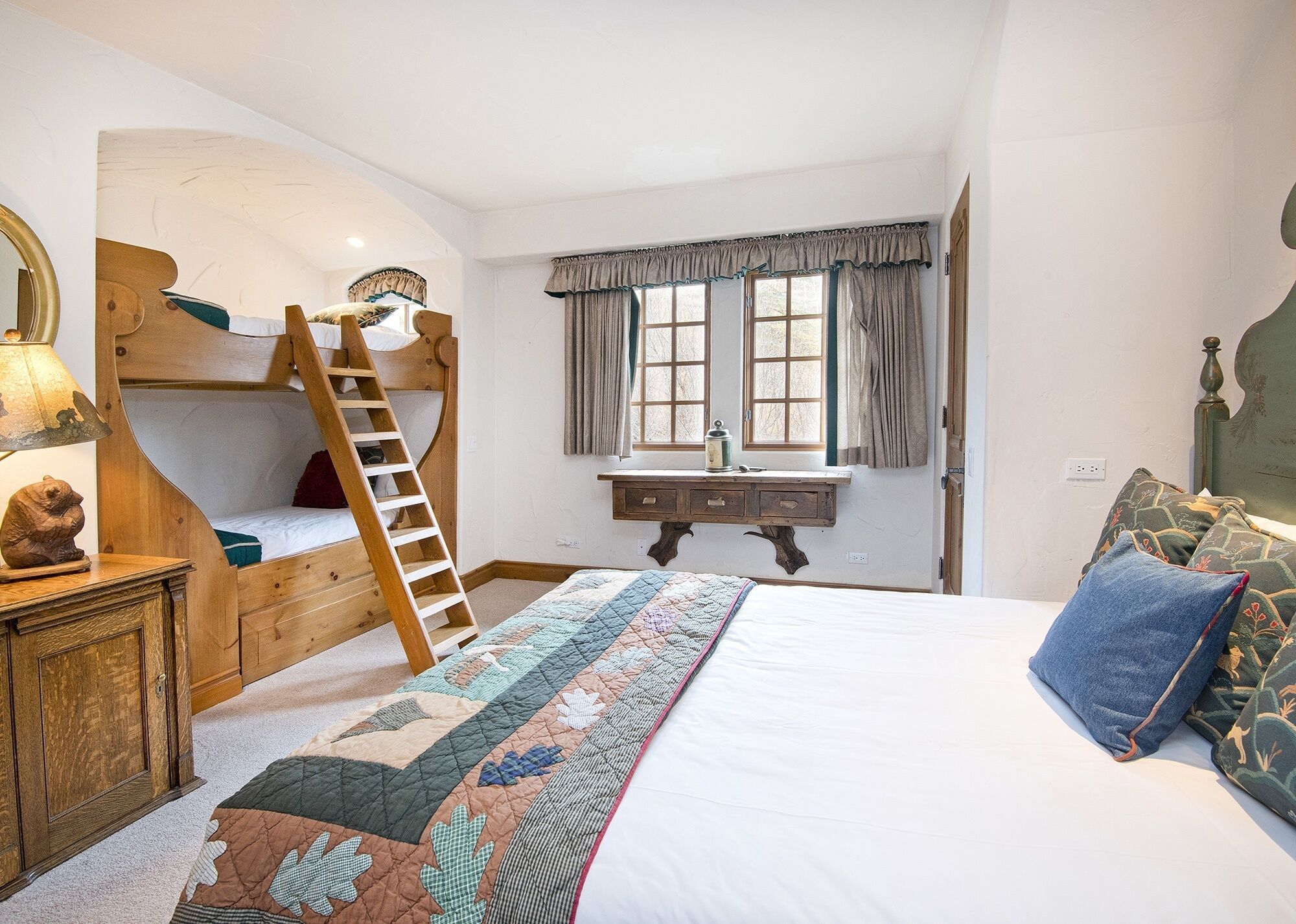 Ein Schlafzimmer mit Etagenbett, Schreibtisch mit Spiegel, Fenster mit Vorhängen und einem Bett mit Tagesdecke.