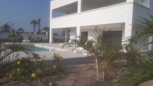 A modern two-story building with a swimming pool, outdoor seating, and tropical plants.