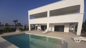 A modern two-story building with a swimming pool, outdoor seating, and a view of palm trees.
