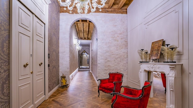 A hallway with wooden parquet flooring, a red upholstered chair, a white fireplace with a mirror, and a door with a brass handle. - Corte Rubbi 6, Deluxe Studio Apartment (Venice)