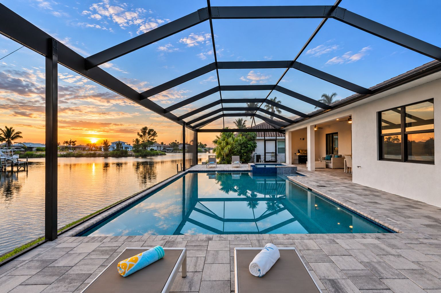 A modern outdoor swimming pool with a glass ceiling, a view of a sunset over water, and a patio area with lounge chairs and a towel.