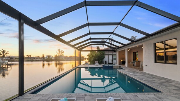 A modern indoor pool with a glass ceiling, a view of a lake, and a patio area with lounge chairs. - Intervillas Florida - Villa Cayman (Cape Coral)
