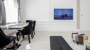 Uma área de jantar moderna com uma mesa branca, cadeiras estofadas pretas e uma televisão de tela plana grande montada na parede, exibindo uma paisagem marítima.
